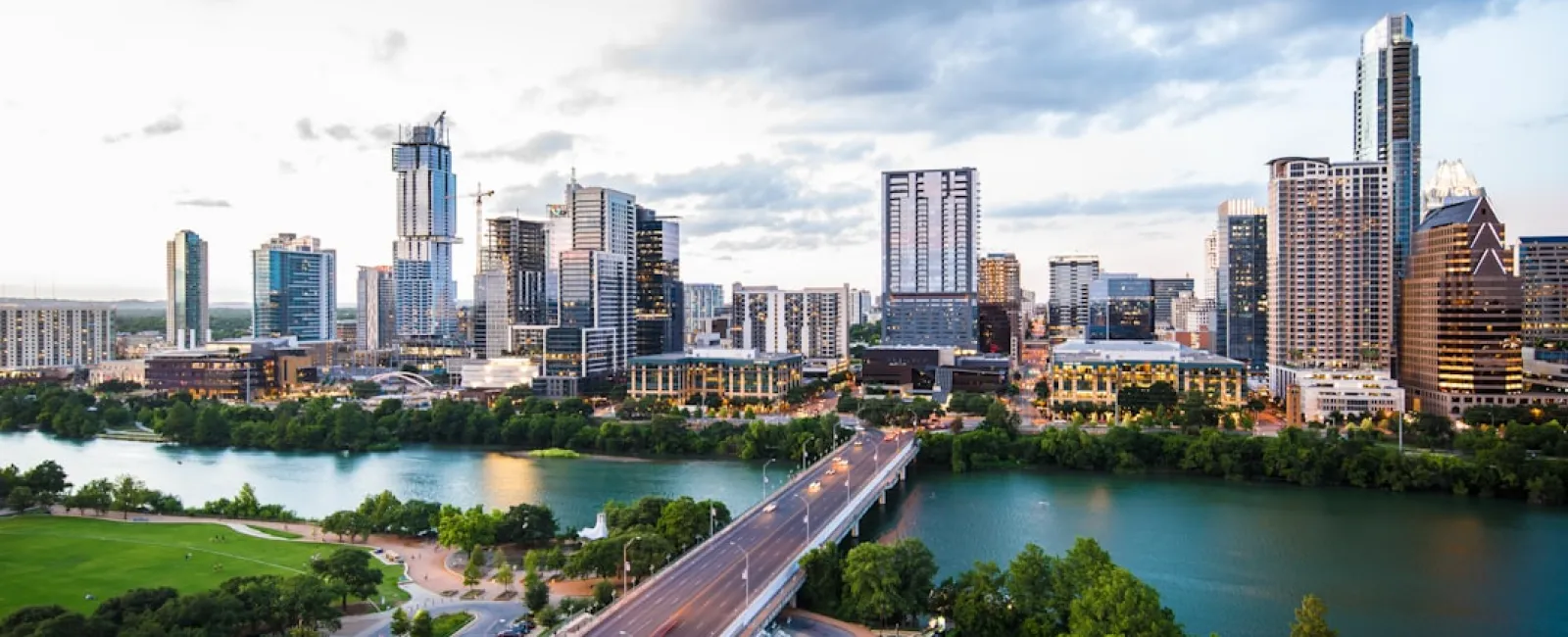 City skyline with tall buildings along a river and a busy bridge during early evening with partly cloudy sky.