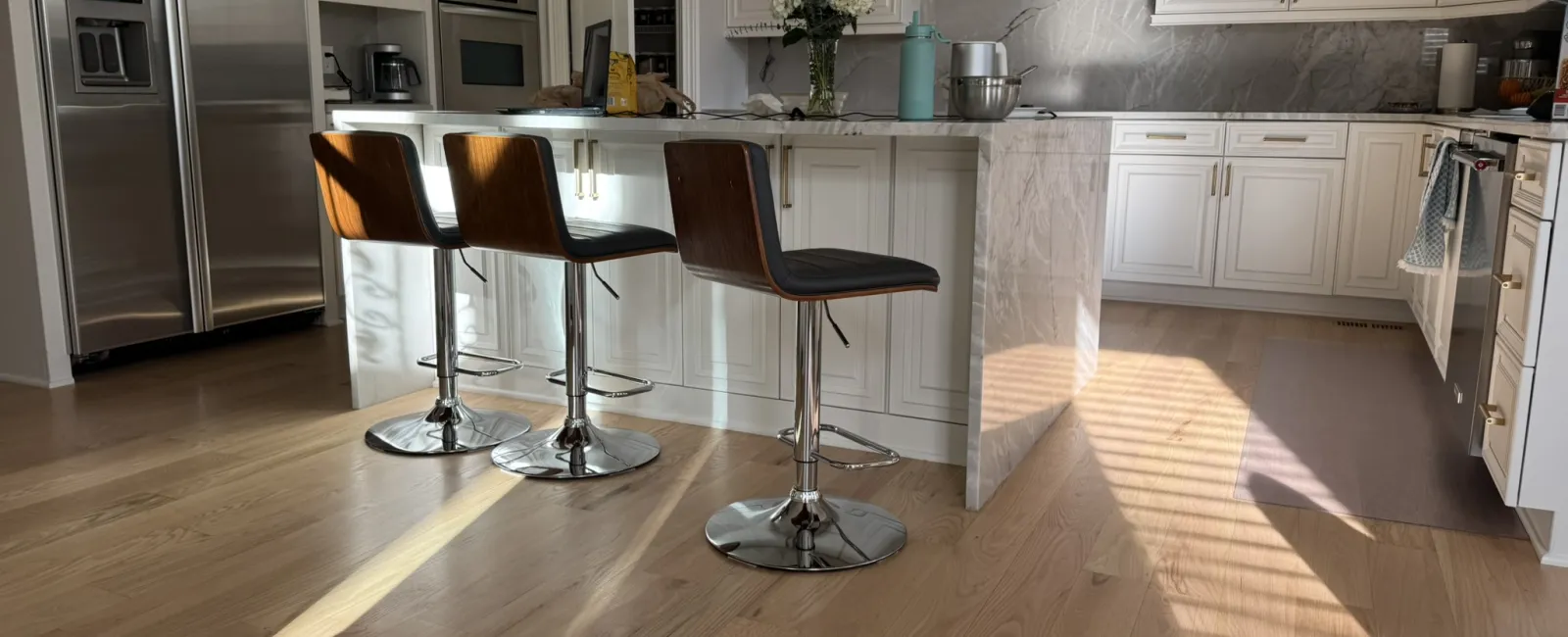 Modern white kitchen with marble island, wooden bar stools, stainless steel appliances, and sunlight on hardwood floor.