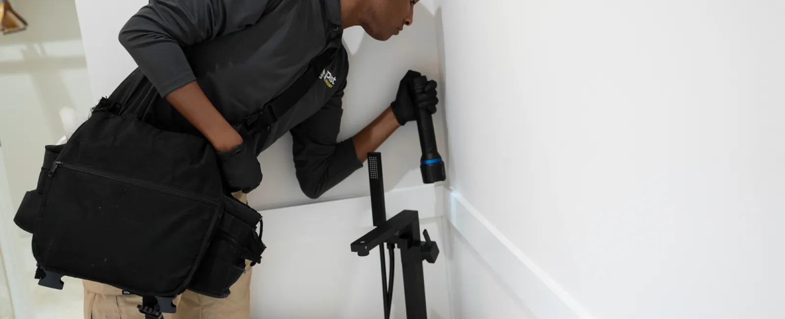 PestMaster technician inspecting for pests in bathroom wall near freestanding tub with flashlight and tool bag.