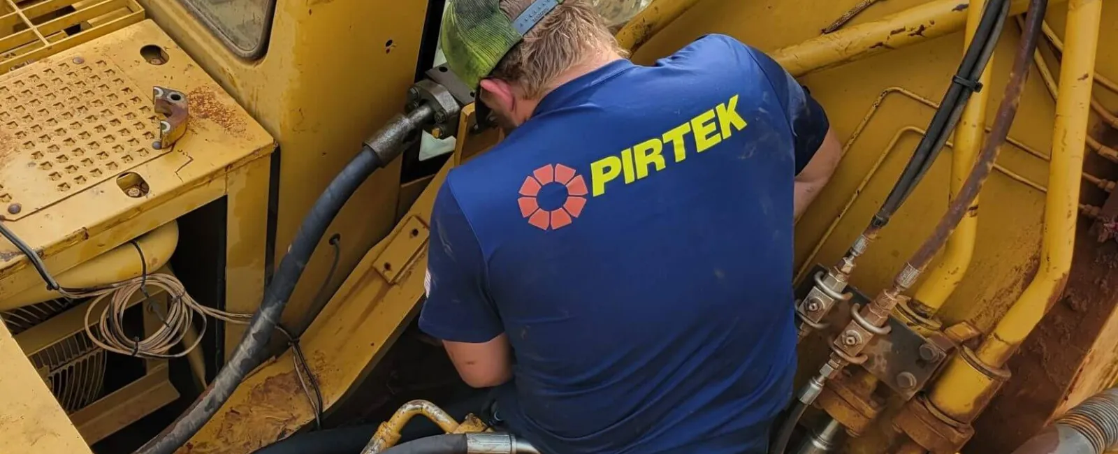 Worker in blue PIRTEK shirt repairs hydraulic machinery on yellow heavy equipment with various hoses and metal parts.