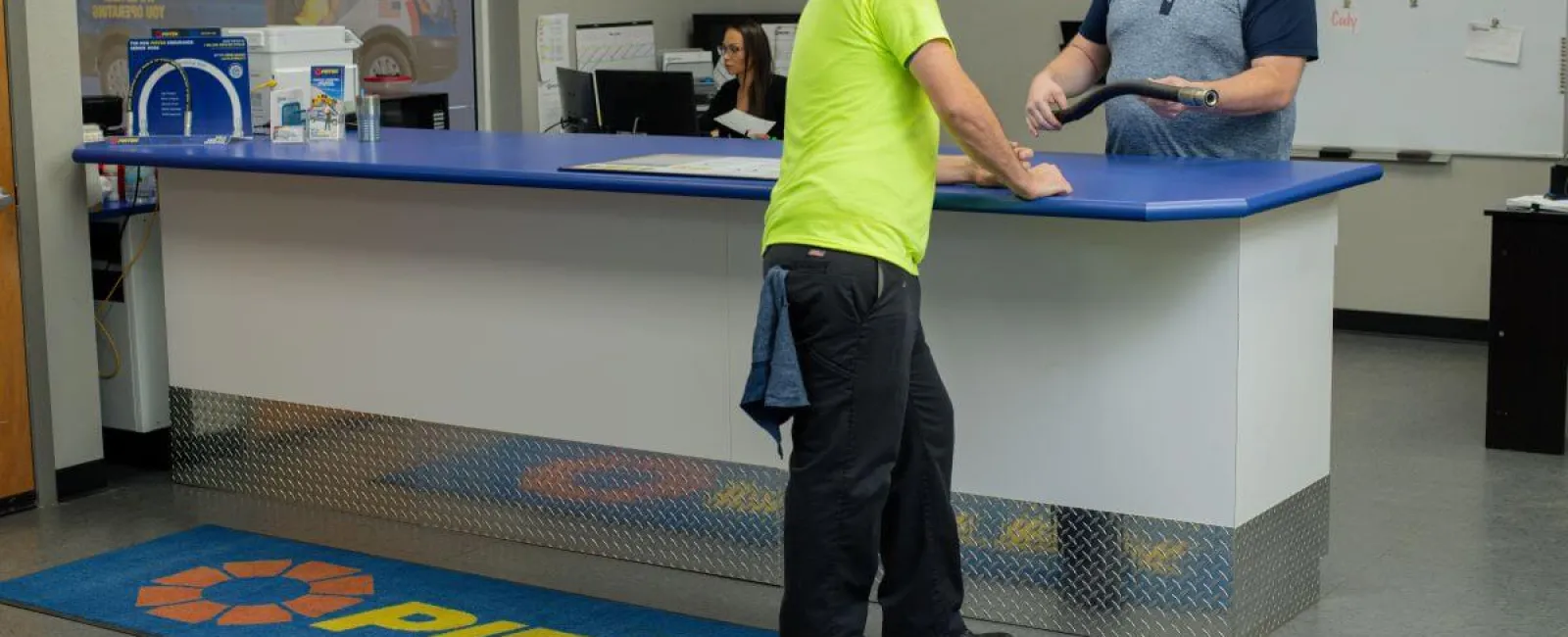 Customer in neon shirt talks to smiling employee at blue counter in Pirtek service center reception area.