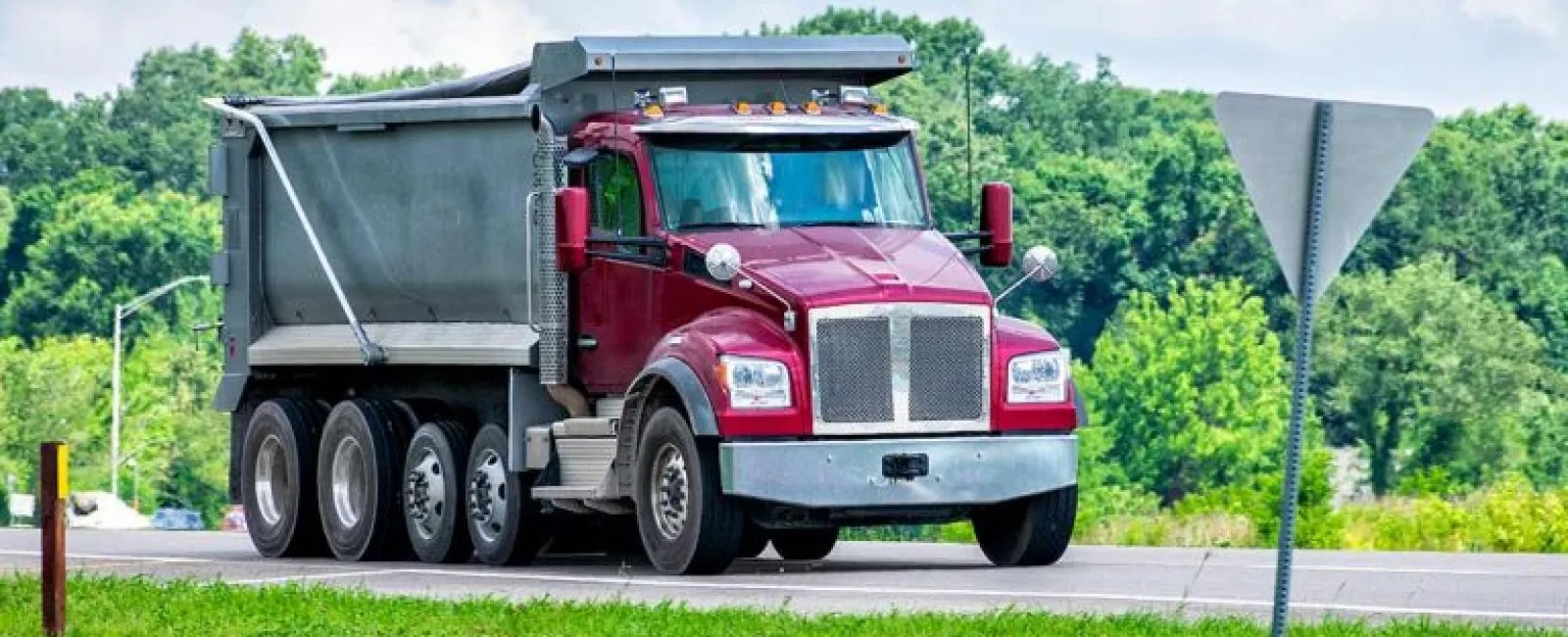 Red dump truck driving on rural road with green trees and blue sky background