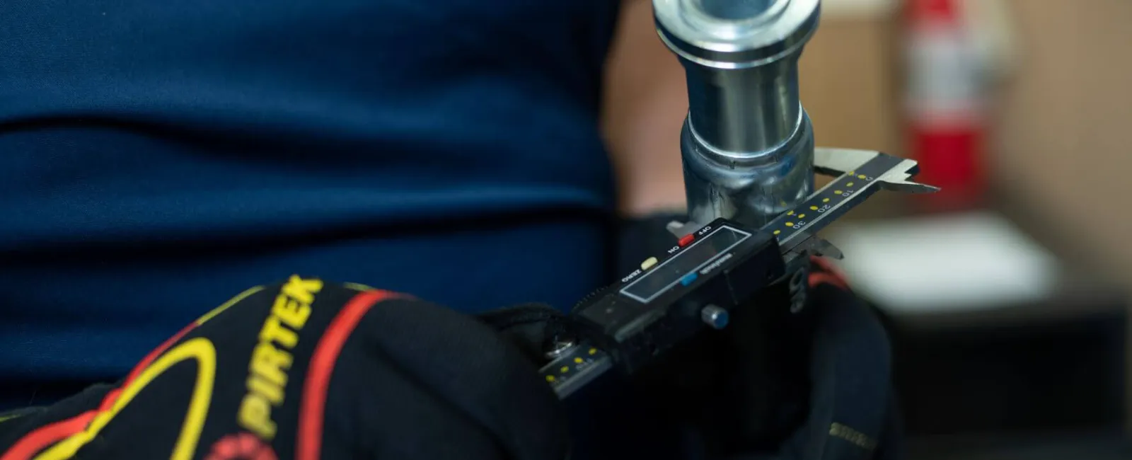 Worker wearing gloves measures metal pipe diameter with digital caliper in workshop setting.