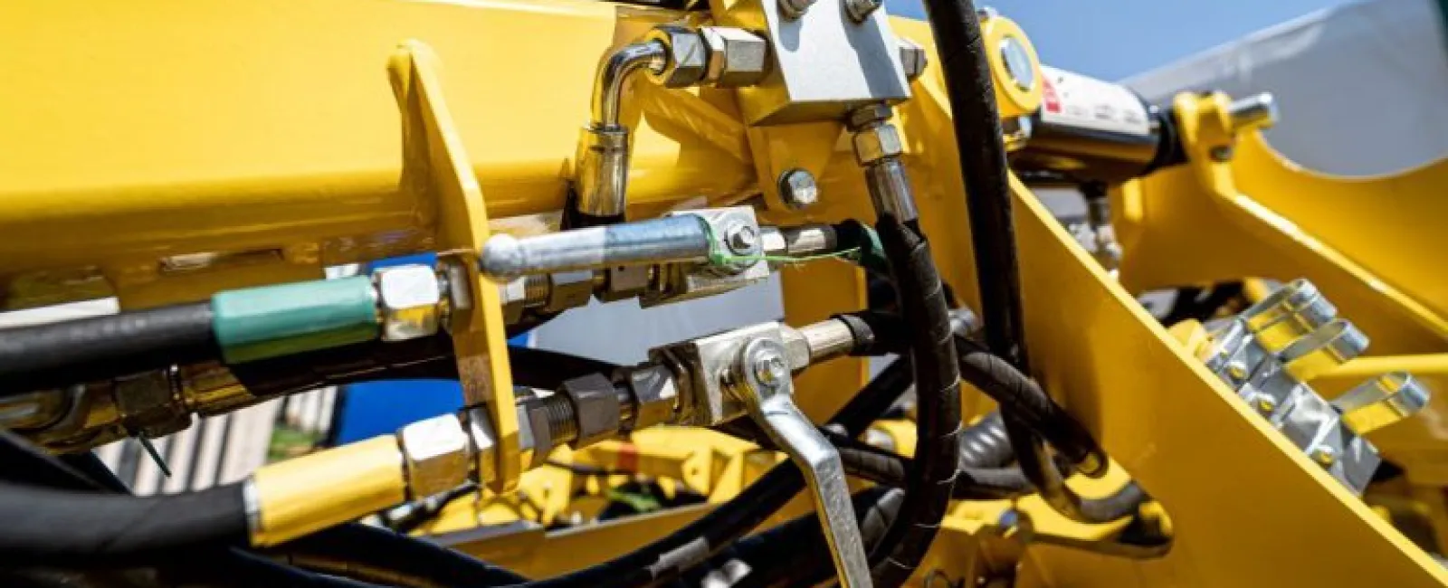 Close-up of yellow heavy machinery hydraulic system with metal pipes, valves, and hoses under bright daylight.