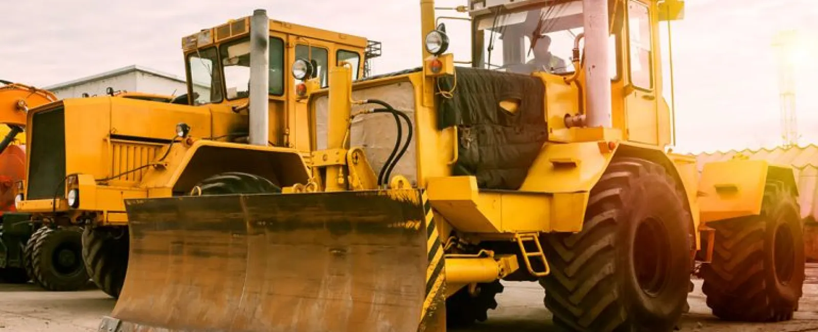 Yellow bulldozers parked on concrete ground under a cloudy sky during sunset at a construction site.