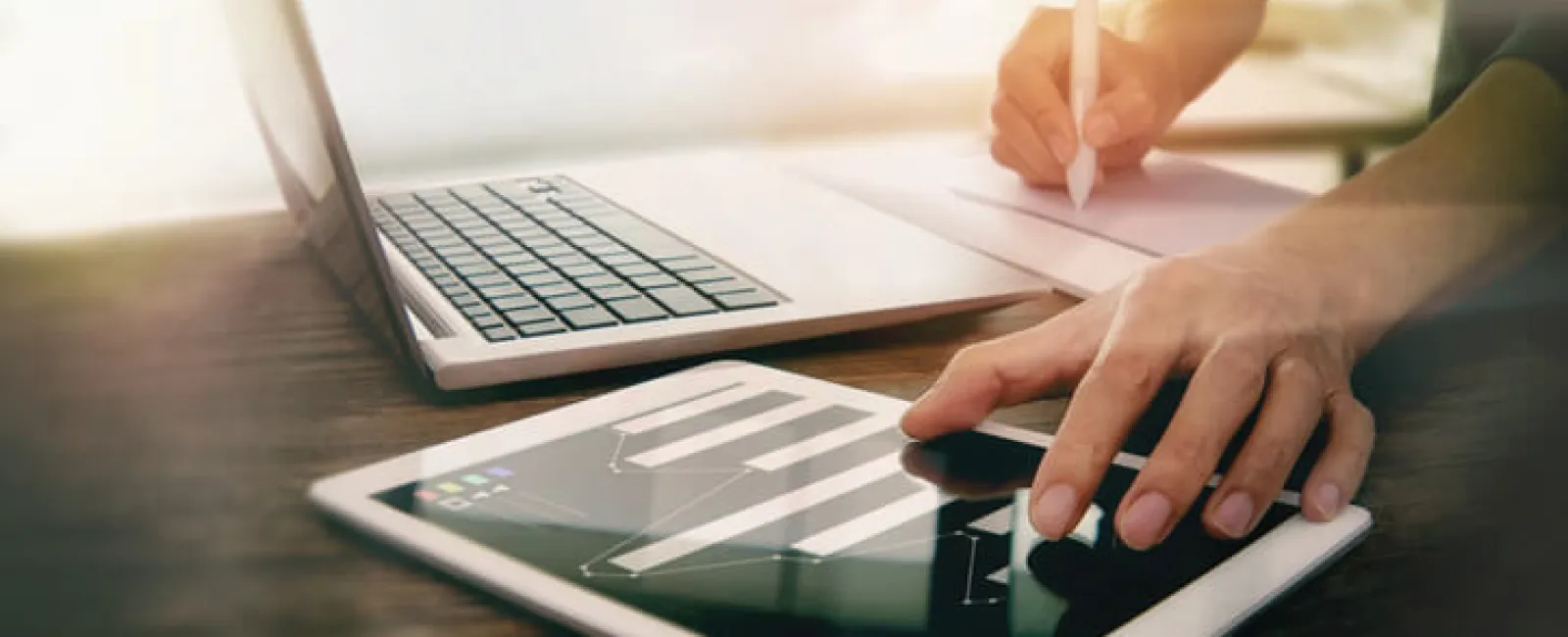 Person analyzing financial data on tablet with bar charts next to laptop and writing notes on paper