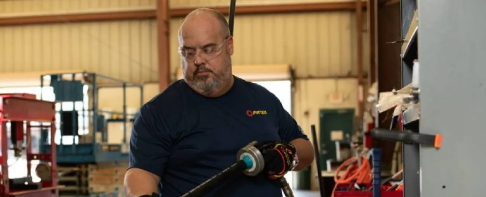 Man in safety glasses and gloves using a power tool on a metal pipe inside an industrial workshop.