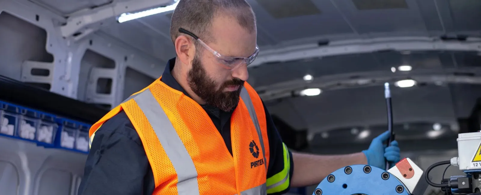 Technician wearing orange safety vest and gloves operating blue hydraulic equipment inside a van workshop.