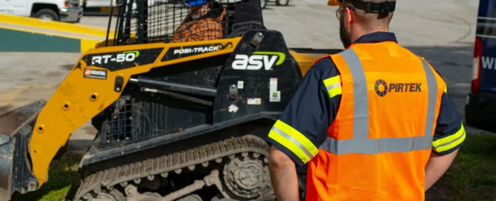 Construction worker in a Pirtek safety vest and helmet observing a yellow ASV Posi-Track RT-50 skid steer loader.