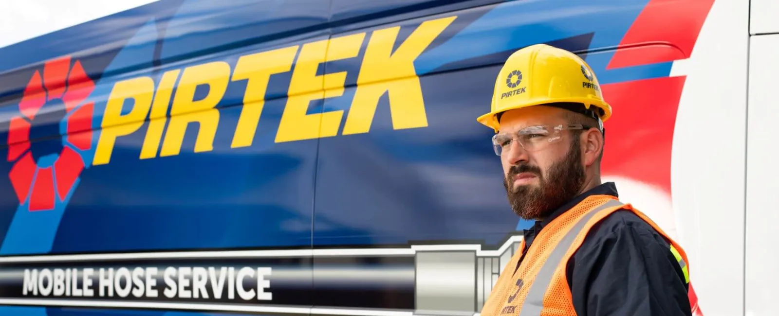 Worker in safety gear using a device in front of a Pirtek mobile hose service van with branding and slogan visible.