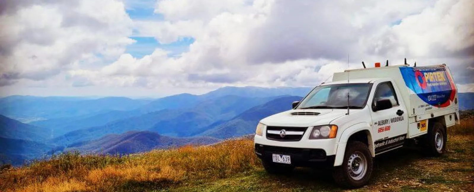 White utility truck parked on grassy hilltop with mountains and cloudy sky in the background