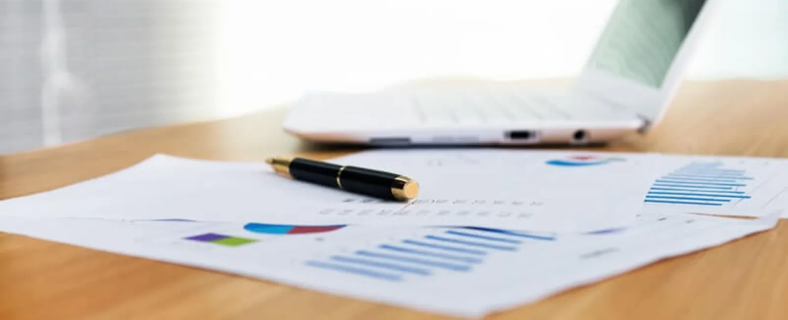 Black pen on financial charts and graphs with laptop on a wooden desk in a bright office.