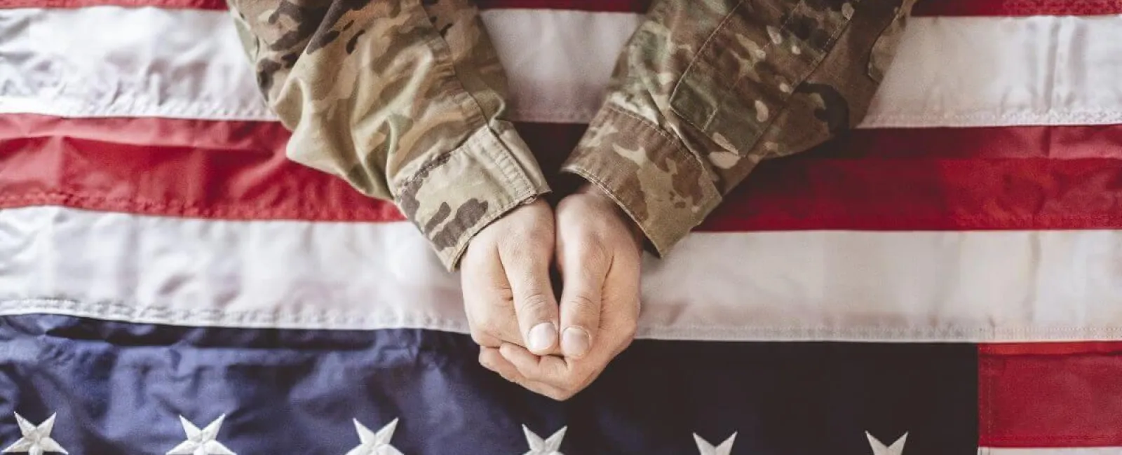 Hands of a soldier in camouflage uniform resting on a folded American flag.