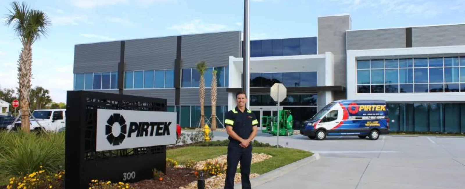 Man in uniform standing outside modern Pirtek building with company van and American flag visible.