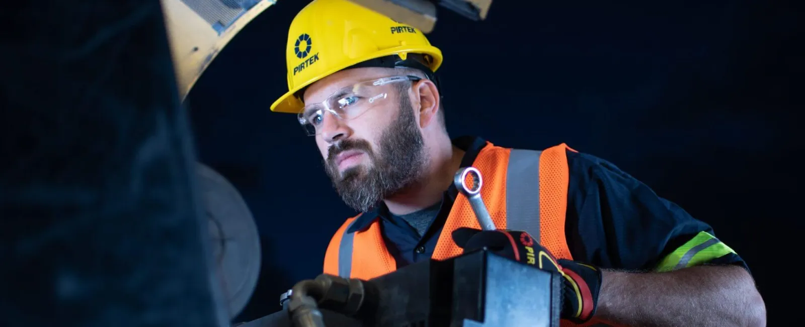 Worker wearing safety gear inspects machinery using a wrench under dim lighting conditions.