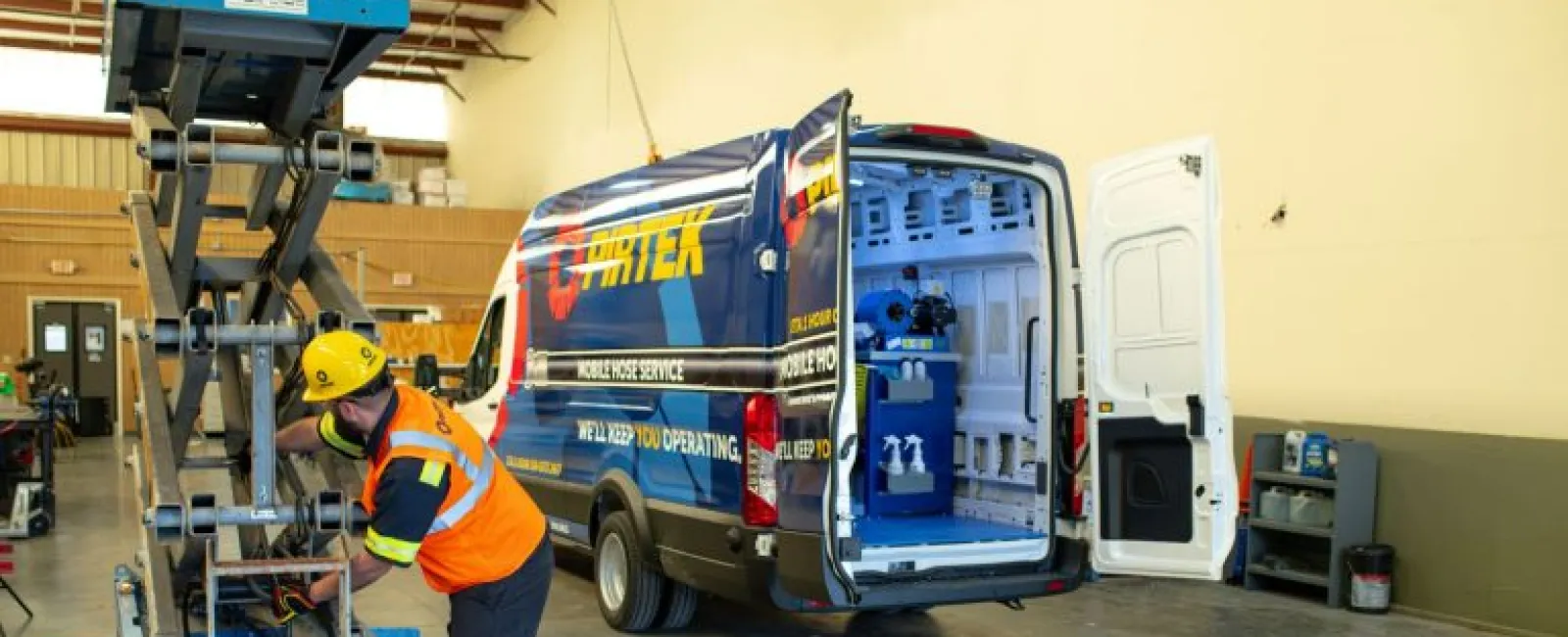 Technician in safety gear working on lift equipment next to a service van inside a spacious workshop.