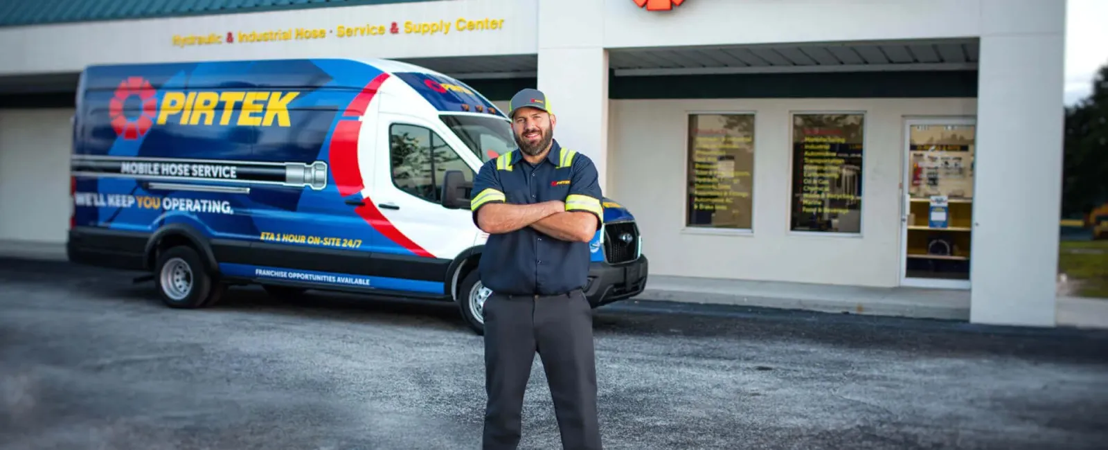 Technician standing confidently with arms crossed in front of a Pirtek service van and store entrance.