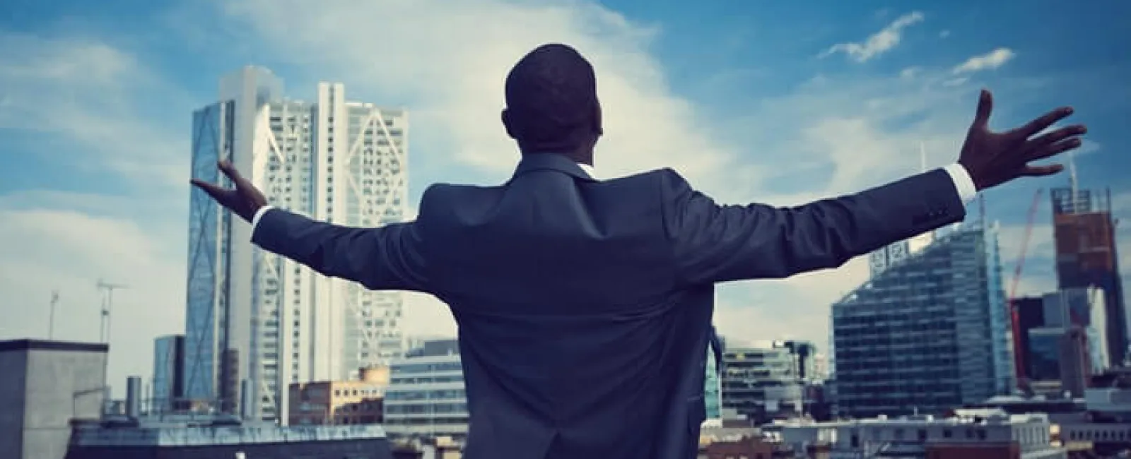 Businessman in suit stands on rooftop with arms outstretched overlooking city skyline under blue sky.