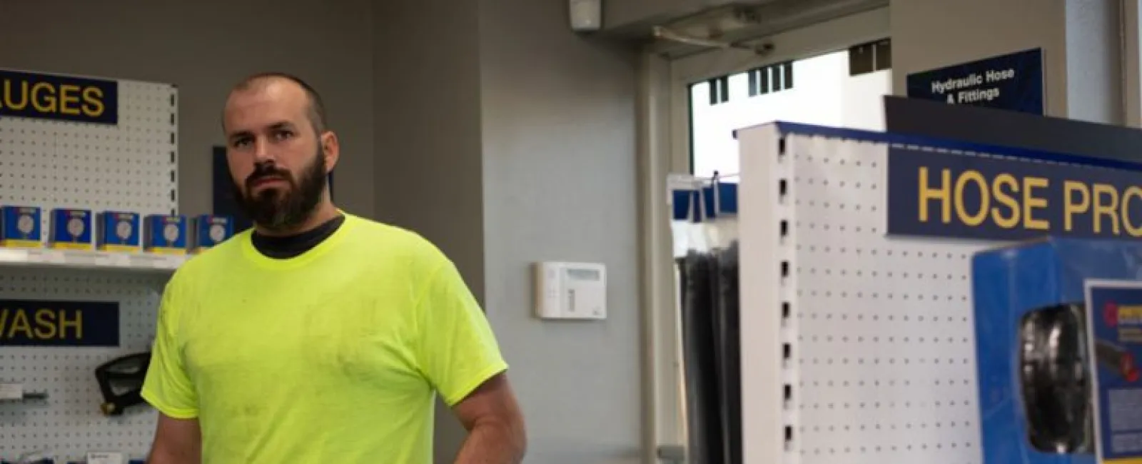 Man in a neon yellow shirt holding a hose in a hardware store aisle with hose products on shelves.