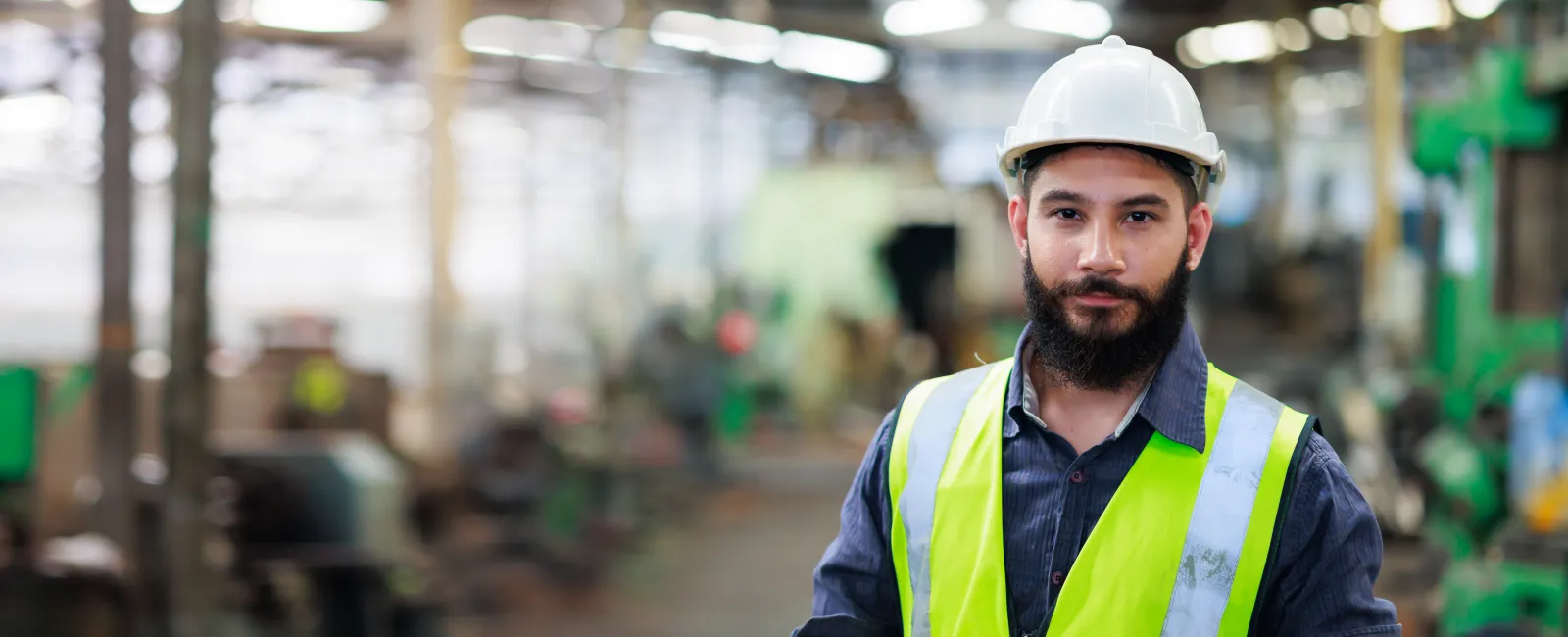 Confident male engineer in hard hat and reflective vest standing inside a factory or industrial setting.