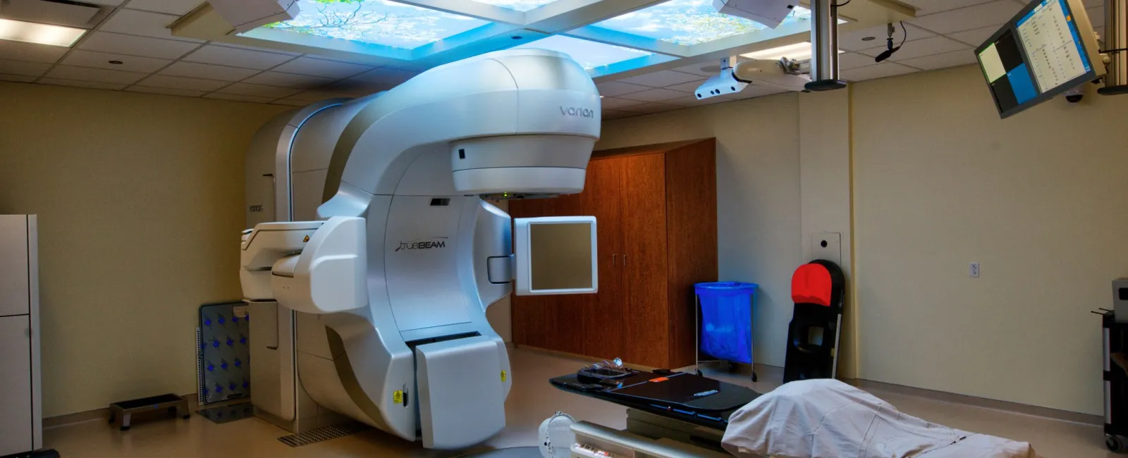 Modern radiation therapy machine in a medical treatment room with illuminated ceiling panels and patient bed.