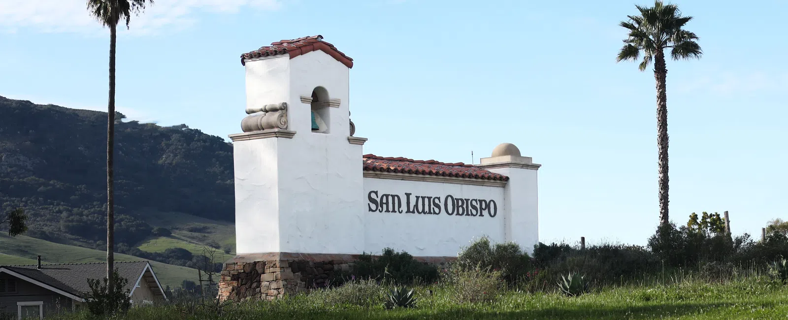 White stucco San Luis Obispo welcome sign with red tile roof and palm trees on grassy hill under blue sky.