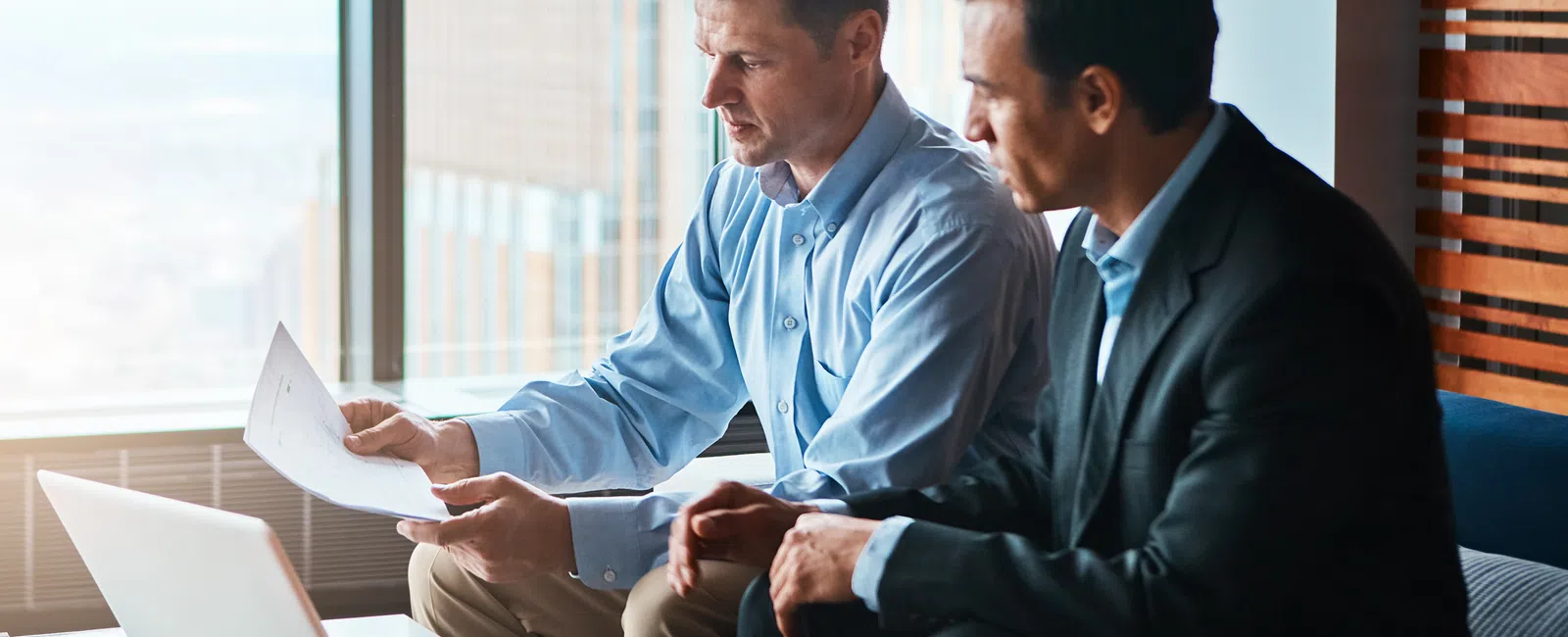 Two businessmen reviewing documents and laptop together in a modern office with city views.