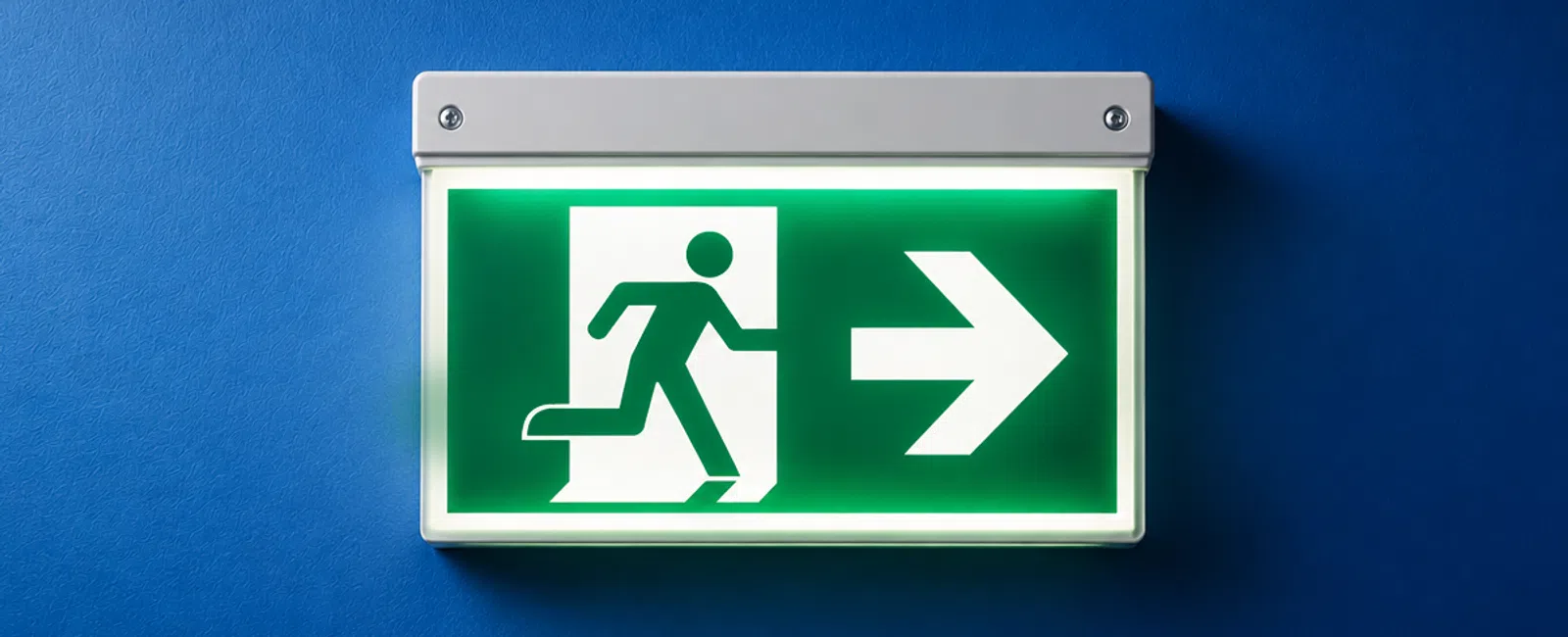 Illuminated emergency exit sign with running figure and arrow pointing right on a blue wall.