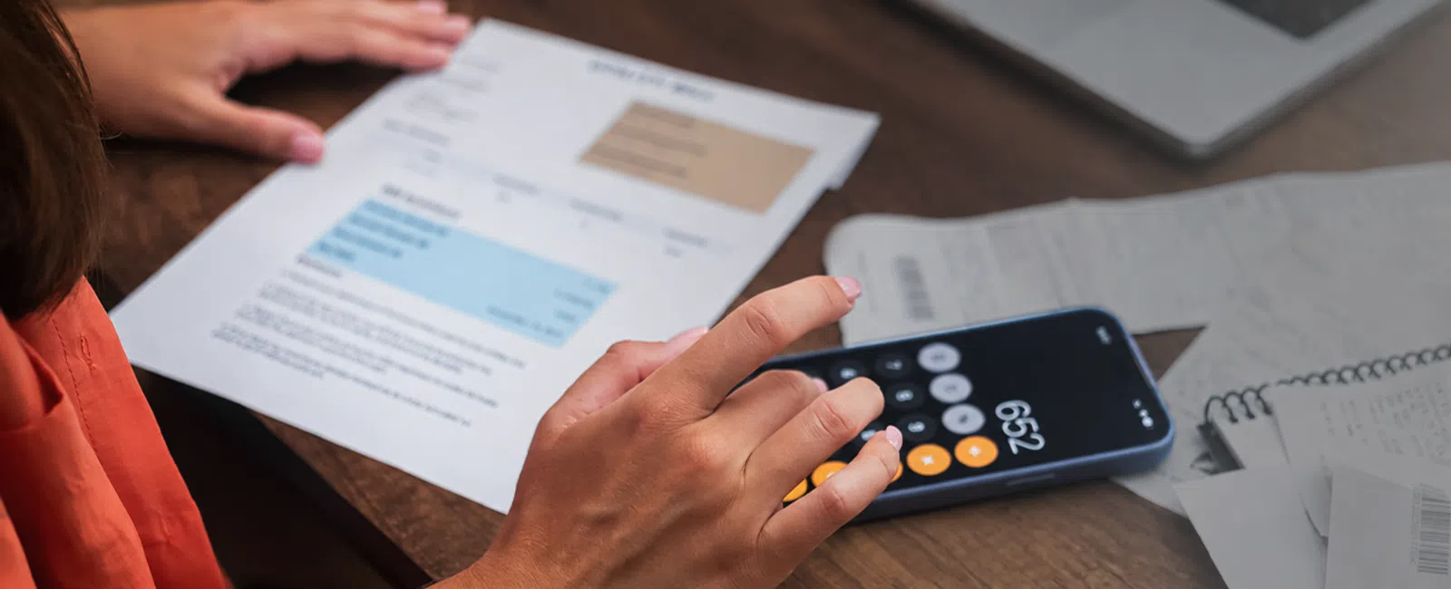 Person using smartphone calculator and reviewing utility bills on wooden desk with laptop and receipts nearby