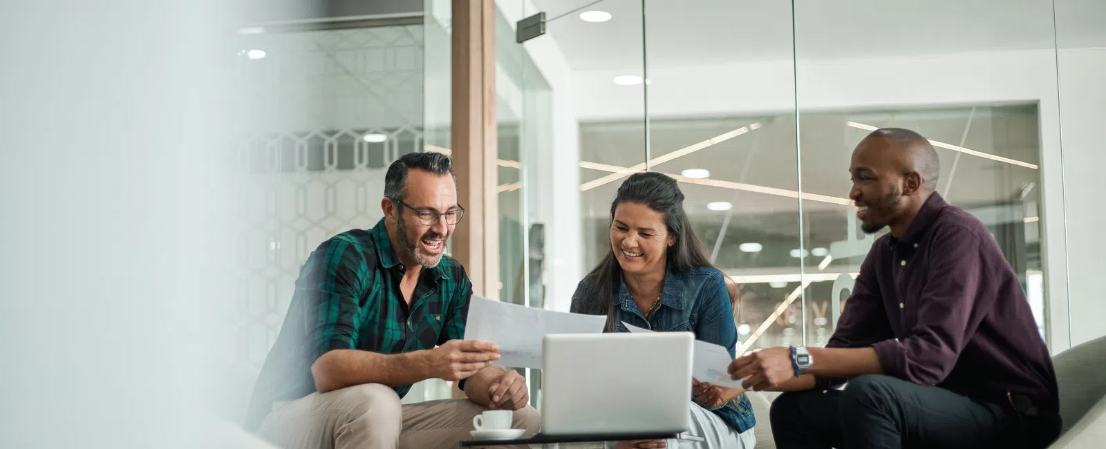 Three colleagues smiling and discussing documents around a laptop in a modern office meeting area.