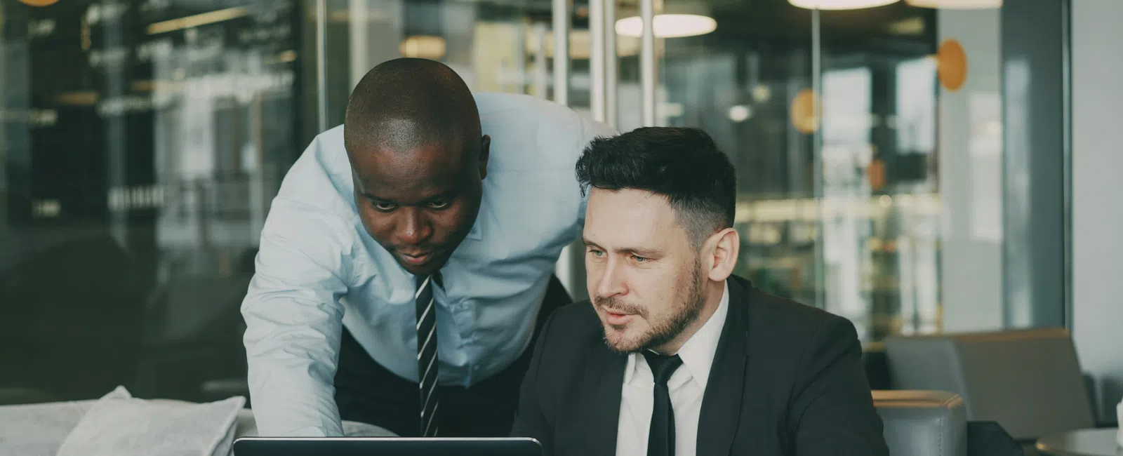 Two businessmen in office attire collaborating over a laptop with a coffee cup on the table nearby