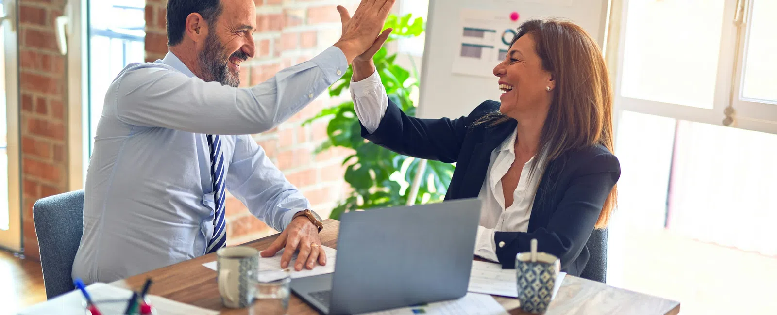 Two professional colleagues sharing a high five over a desk with laptop and paperwork in a bright office