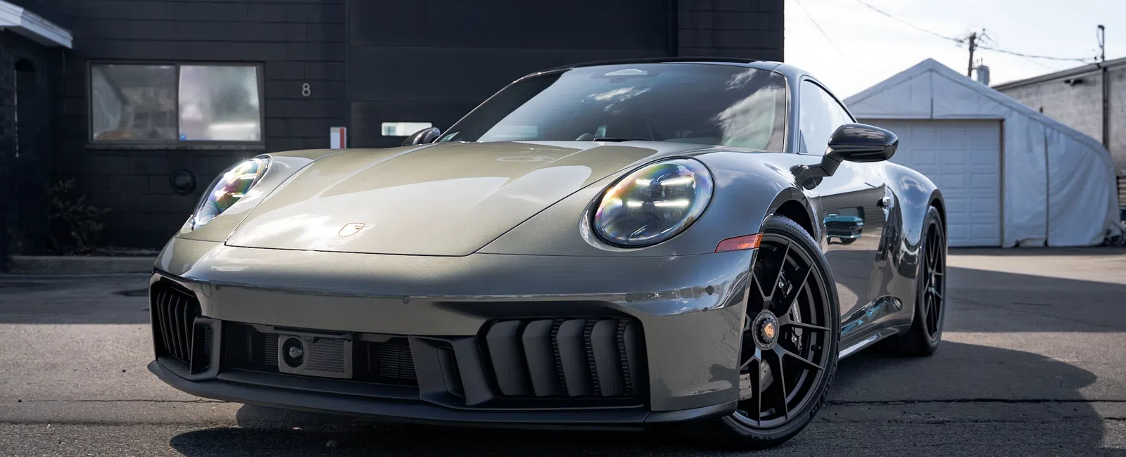 Silver Porsche 911 sports car parked outside a garage under clear sky, showcasing sleek design and black alloy wheels