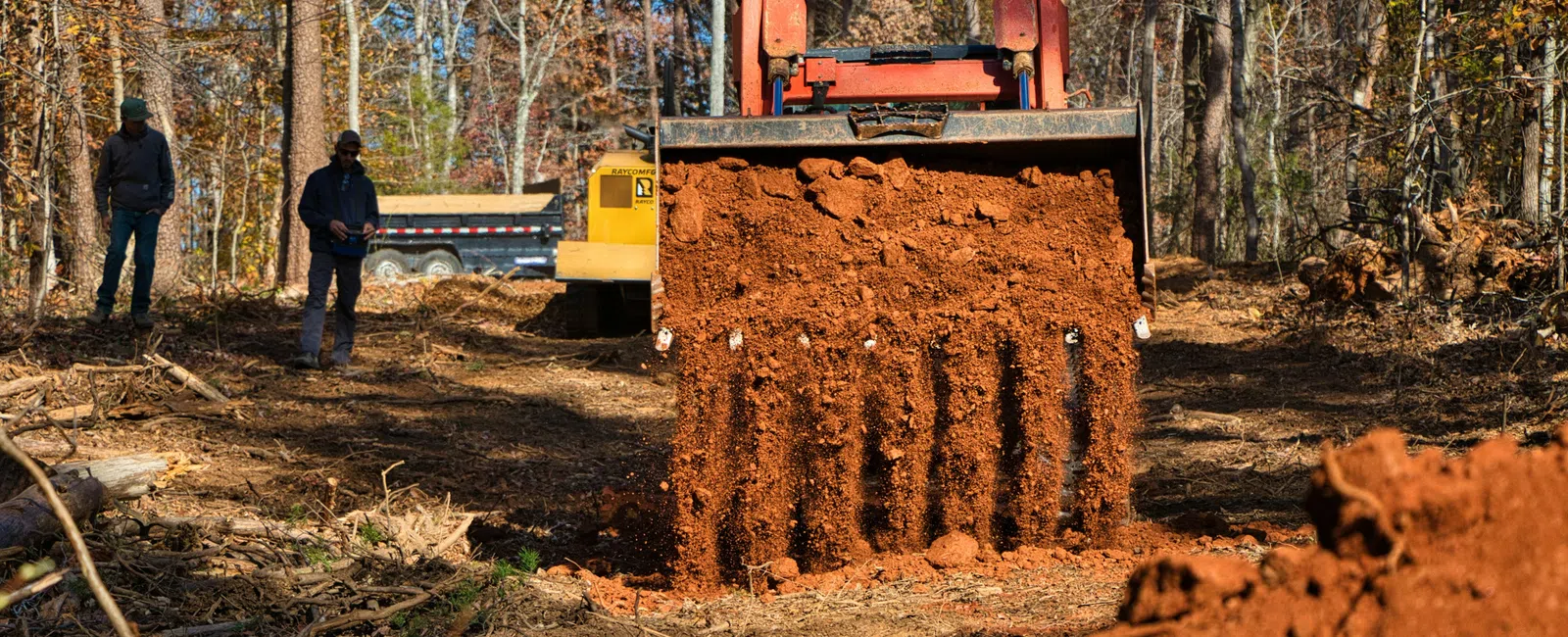 Bulldozer moving red soil in forest clearing with two workers and a dump truck in background on a sunny day