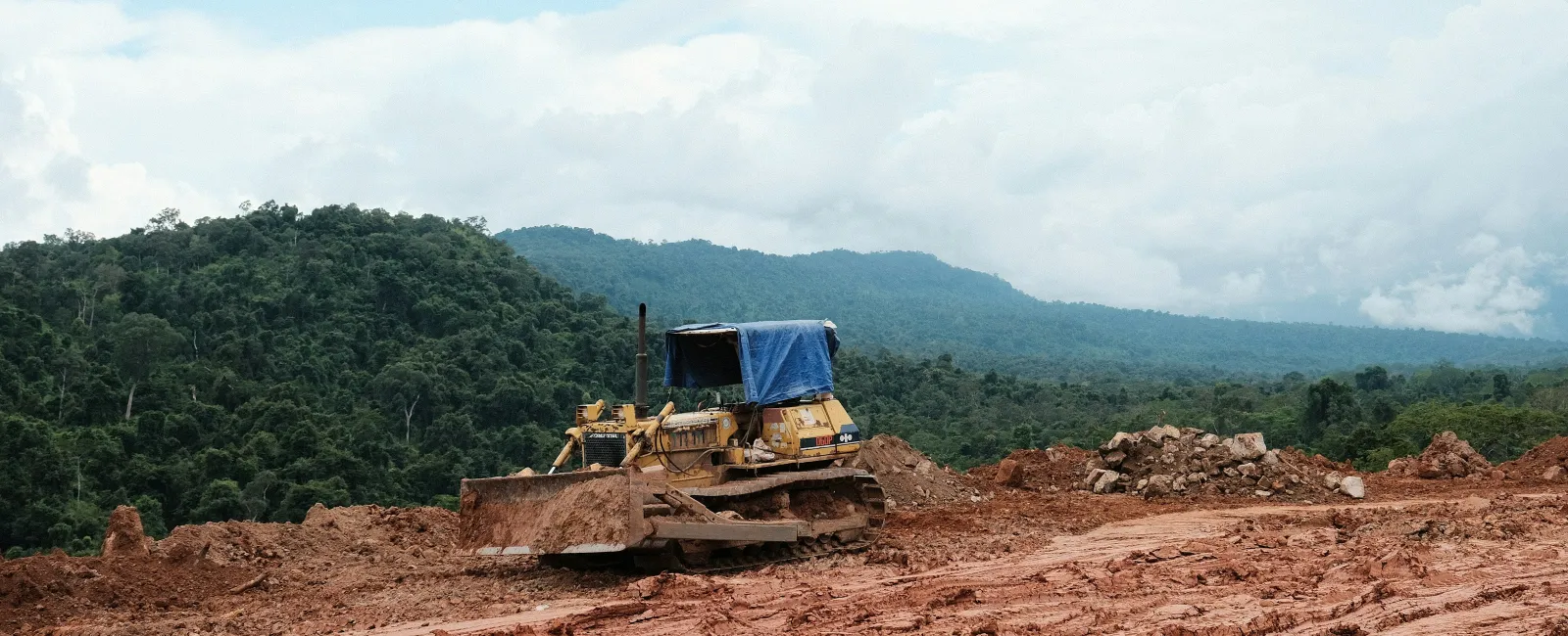 Bulldozer working on cleared red dirt land with forested hills and cloudy sky in the background.