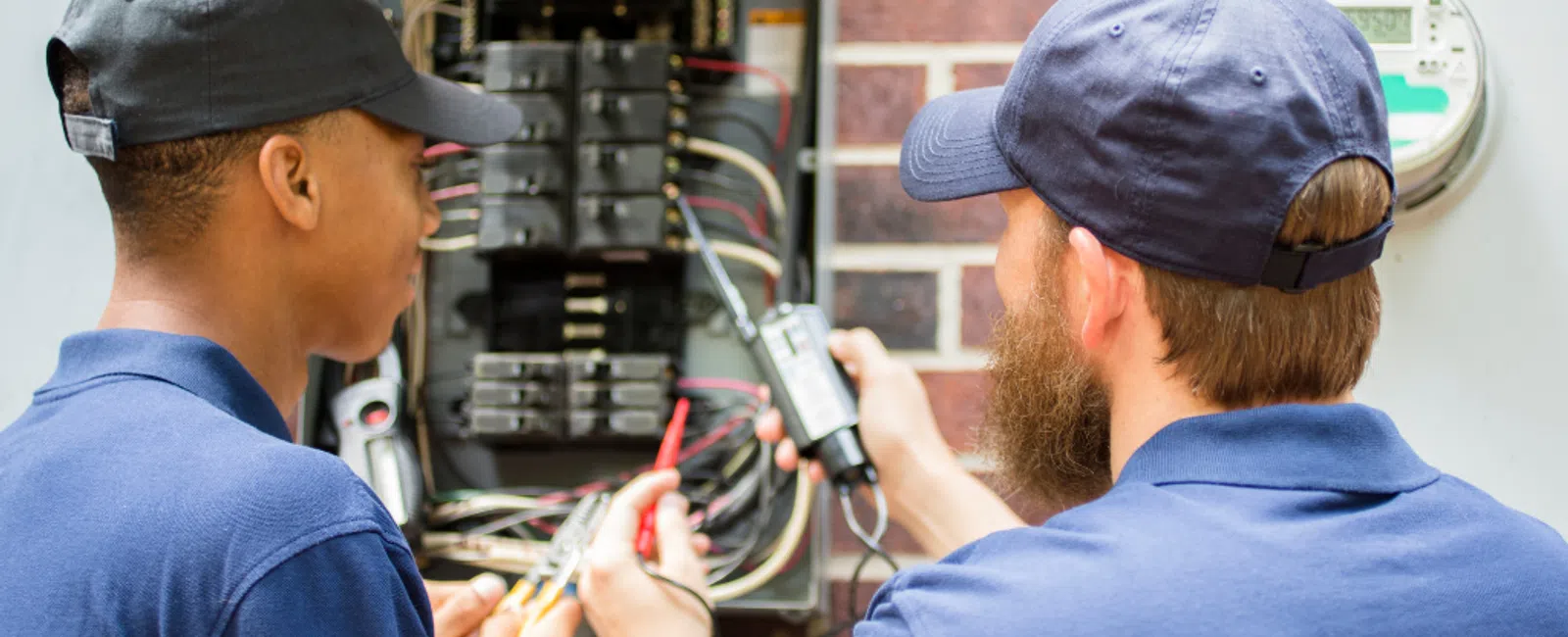 Two electricians working on an electrical panel, testing wires and connections together.