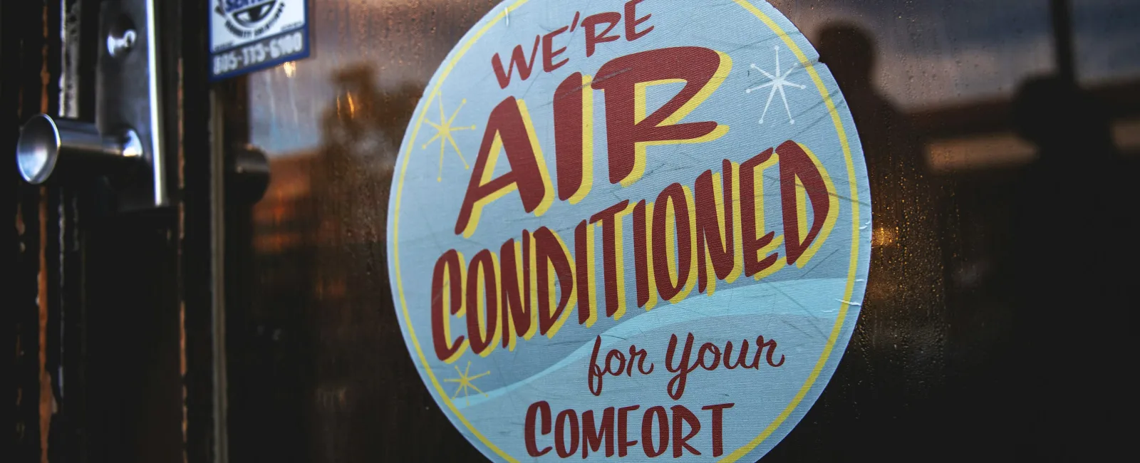 Window sign stating We're Air Conditioned for Your Comfort on a glass door with reflections in warm light