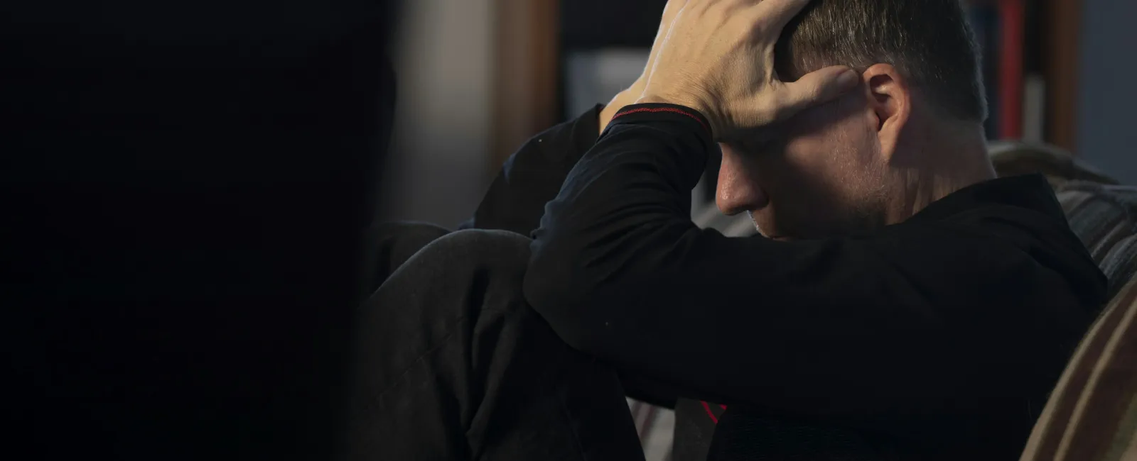 Man sitting on couch with hands on head appearing stressed or anxious in dimly lit room.