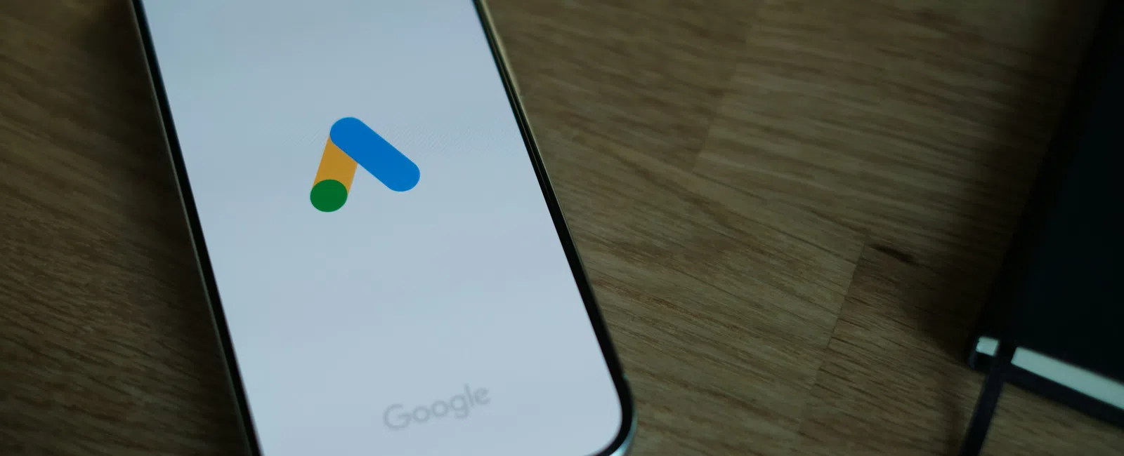 Smartphone displaying the Google Ads logo on a wooden table next to a closed notebook.