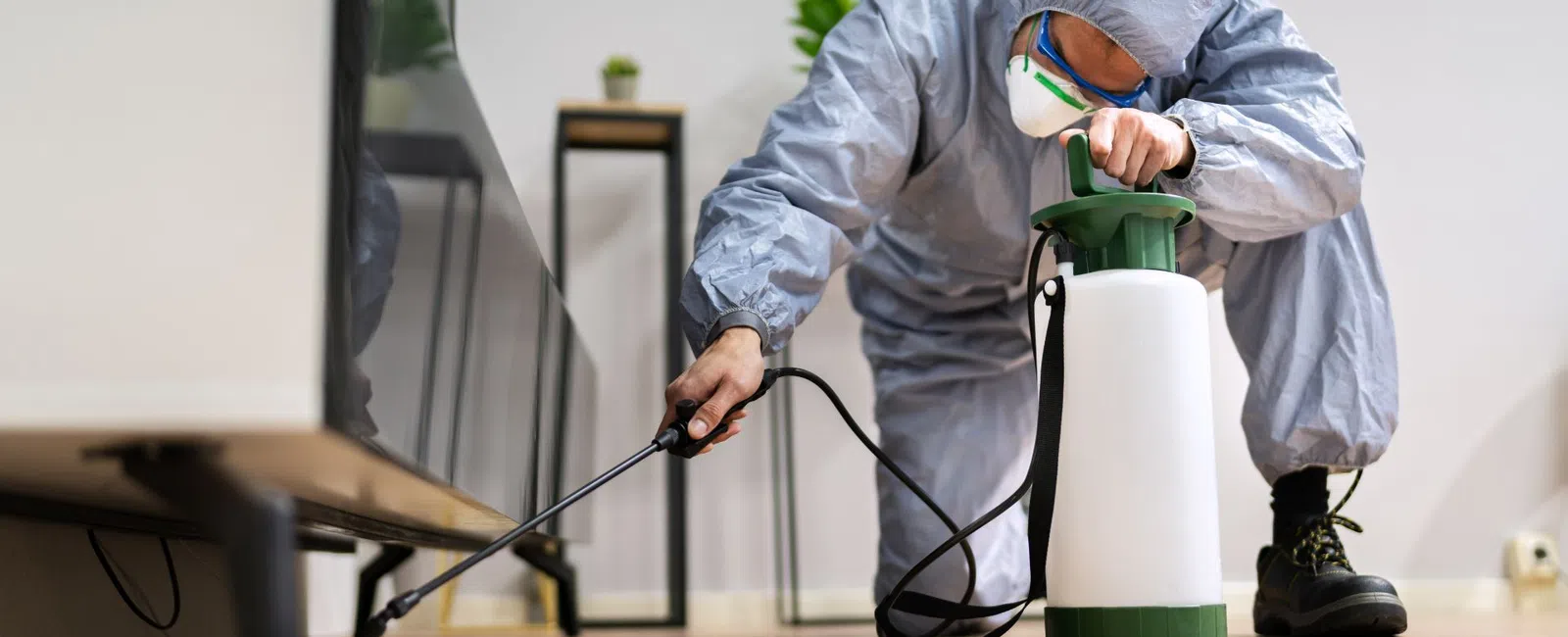 A pest control technician sprays treatment in a living room, wearing protective gear and focused on the task.