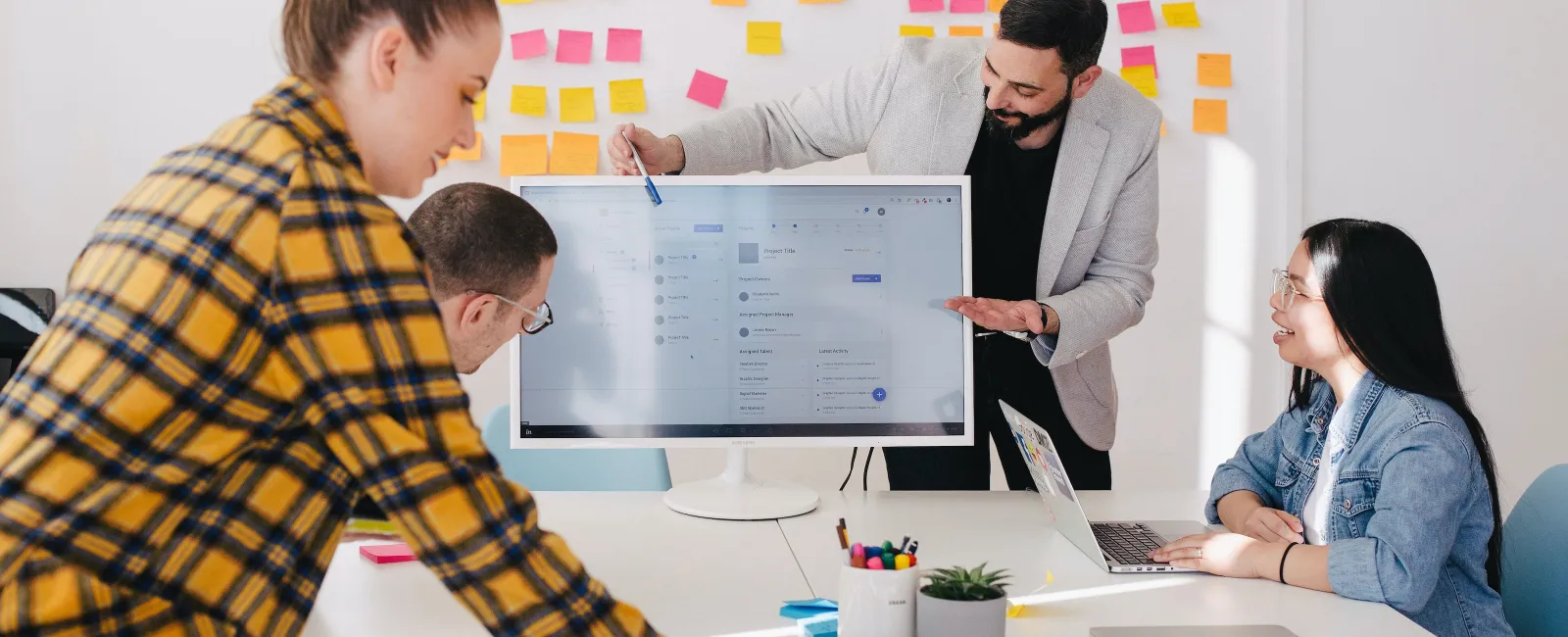 Team members collaborate in office meeting with sticky notes and digital presentation on computer screen.
