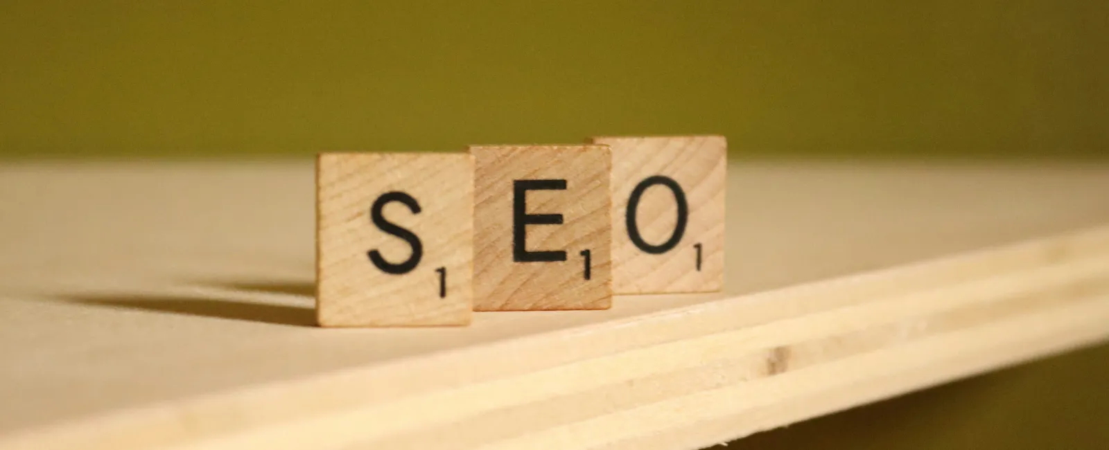 Wooden Scrabble tiles spelling SEO placed on the edge of a light wooden surface with a green background.