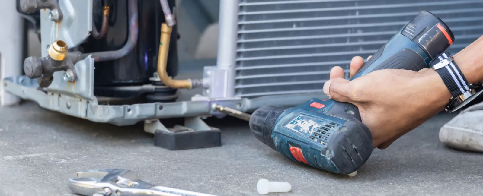 Technician fixing an air conditioning unit using a cordless drill and various hand tools on concrete floor.