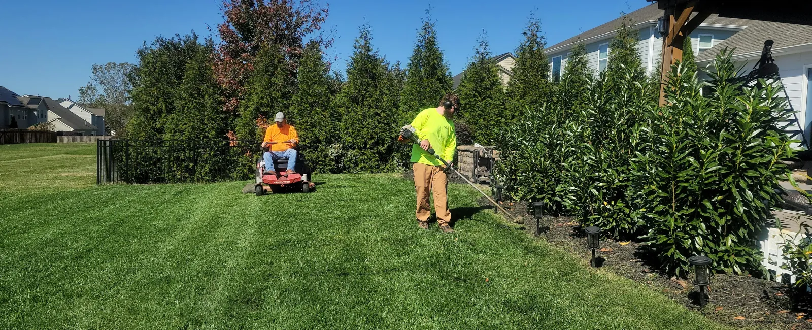 Two landscapers working on lawn care with mower and trimmer in a suburban yard on a sunny day