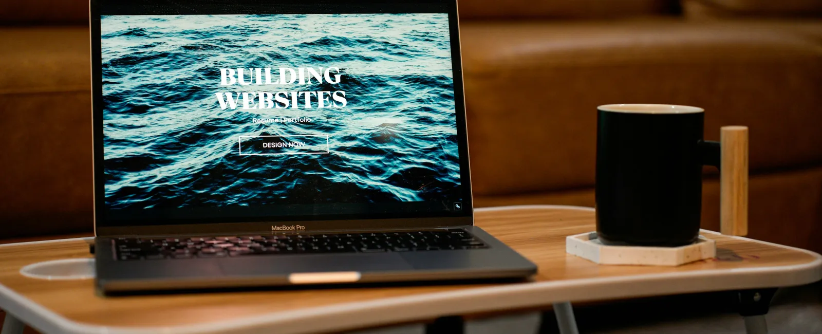 Laptop on wooden table displaying website with ocean waves and text building websites, next to black coffee mug.