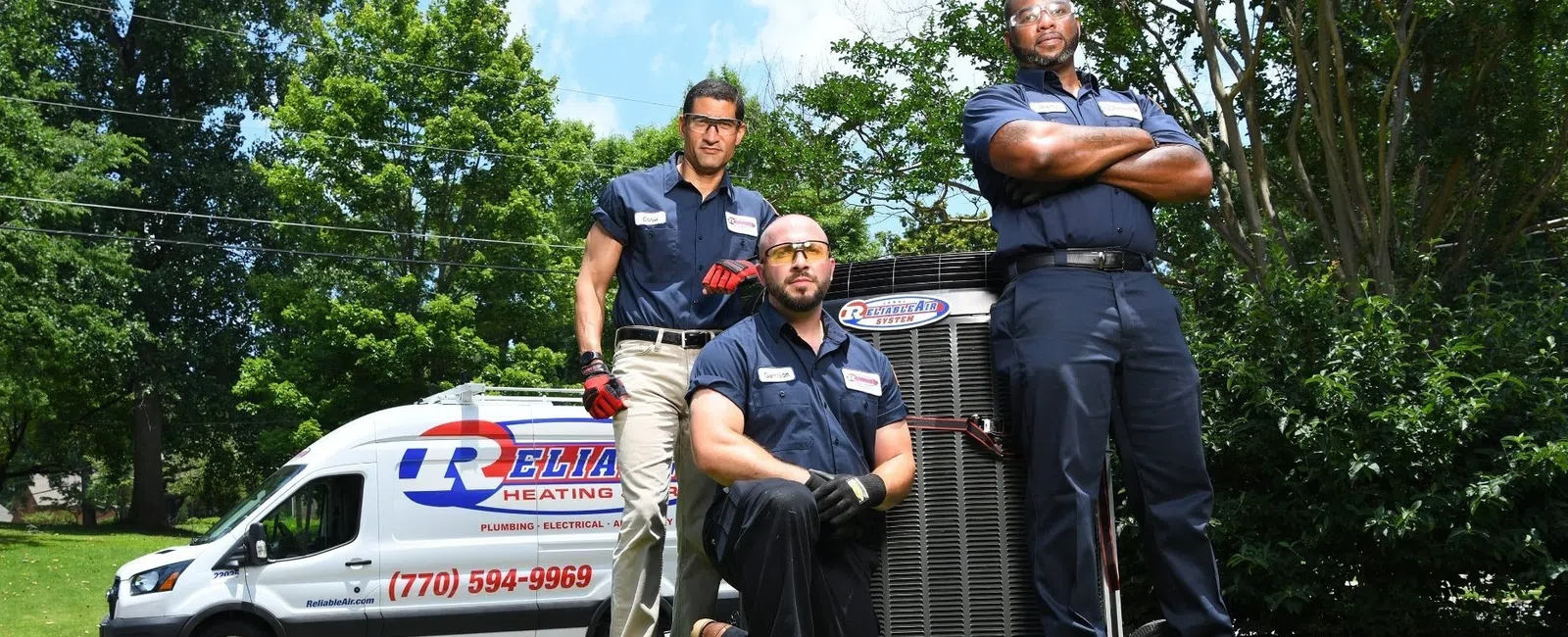 Three HVAC technicians posing with heating unit on green lawn beside company service van on sunny day.