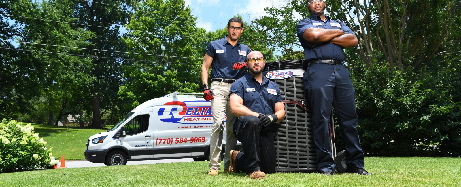 Three HVAC technicians posing with heating unit on green lawn beside company service van on sunny day.