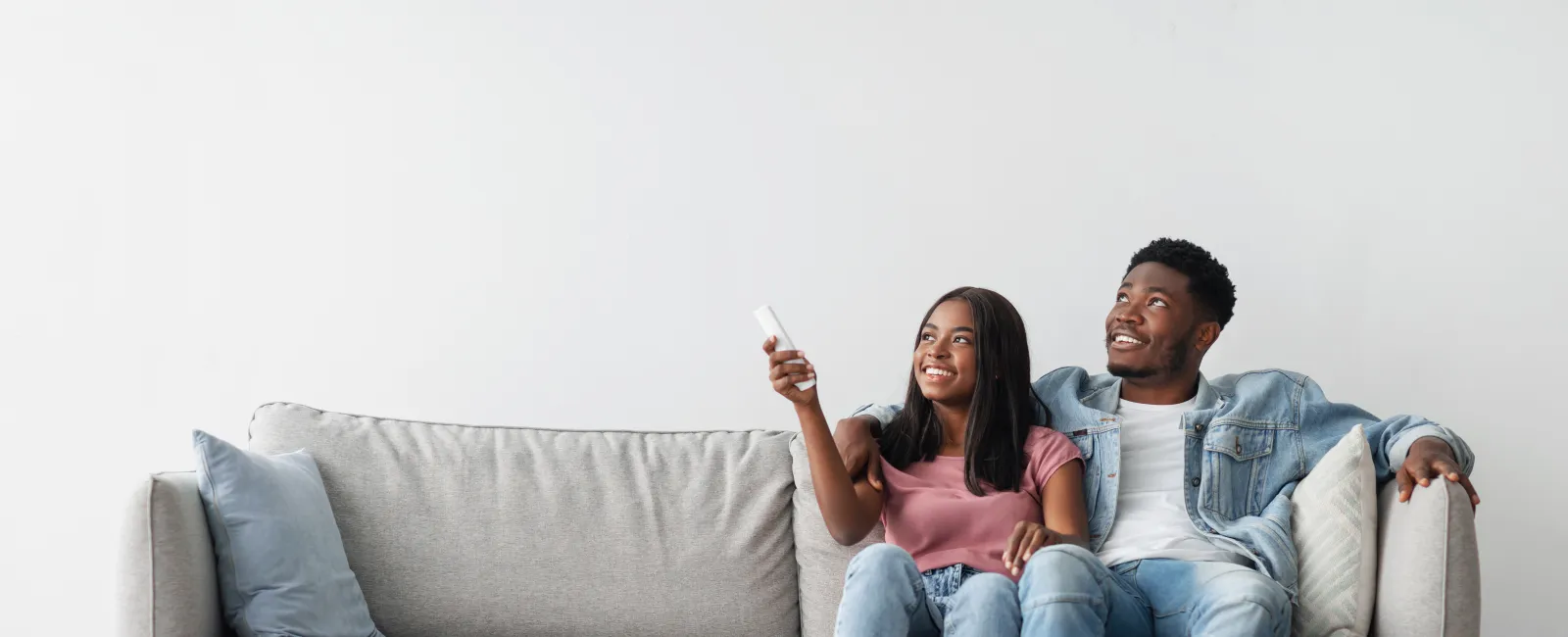 Smiling young couple sitting on a gray couch watching TV with remote control in a modern living room.