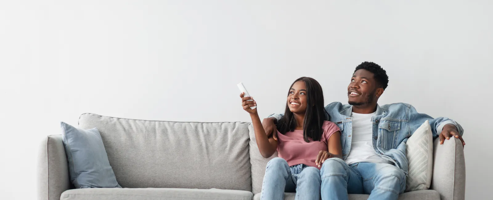 Happy young couple sitting on gray sofa watching TV together with remote control in modern living room