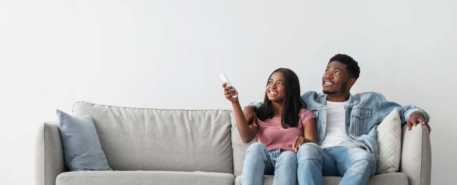 Happy couple sitting on a modern gray sofa watching TV with a remote control in hand in a bright living room.